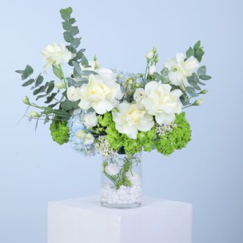 An arrangement of white roses, blue hydrangea, green hydrangea, and lisianthus with eucalyptus leaves in a glass vase.