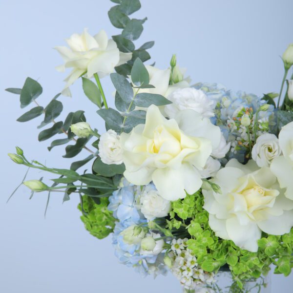 A lovely glass vase filled with white roses, hydrangeas in blue and green, lisianthus, and fresh eucalyptus leaves.