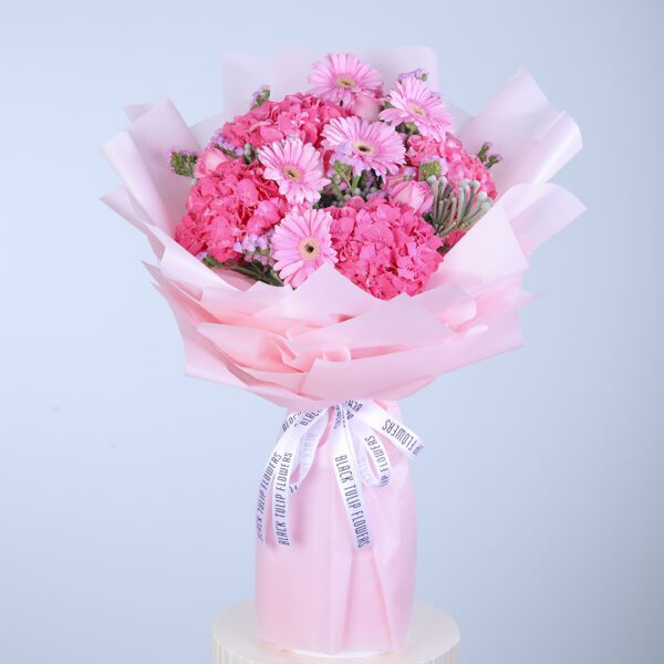 A vibrant pink bouquet featuring gerberas, hydrangeas, roses, and ferns, wrapped in pink tissue paper and tied with white ribbon