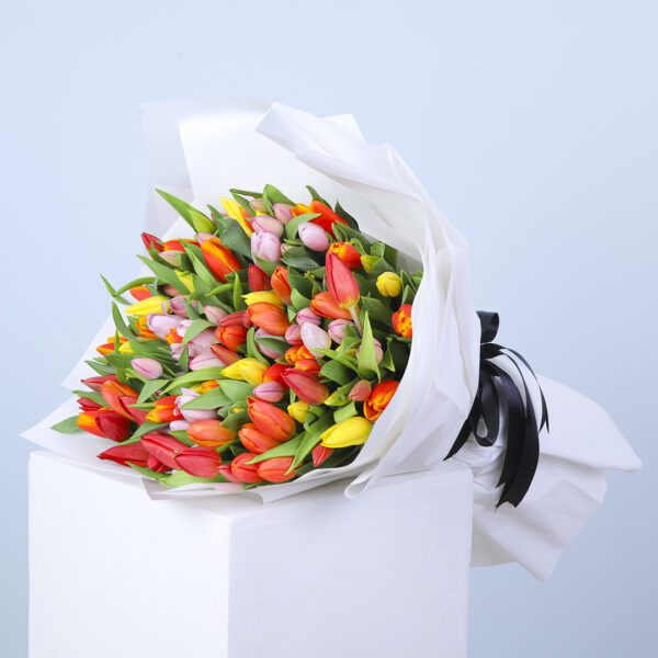 A colorful bouquet of tulips in red, orange, yellow, and pink, wrapped in white paper with a black ribbon