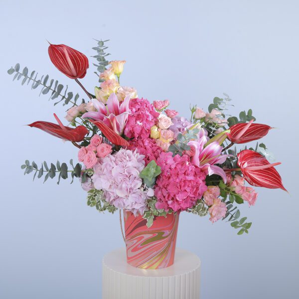 A colorful floral arrangement in a vase, featuring pink hydrangea, red anthurium, roses, lilies, and greenery.