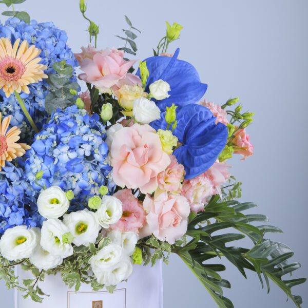 A serene spring bouquet of blue hydrangeas, daisies, and peach and white carnations, nestled in a white box with lush greenery.