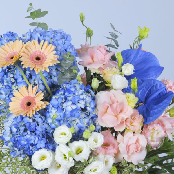 A breathtaking floral masterpiece featuring blue hydrangeas, daisies, and peach and white carnations, elegantly displayed in a white box with vibrant greenery.