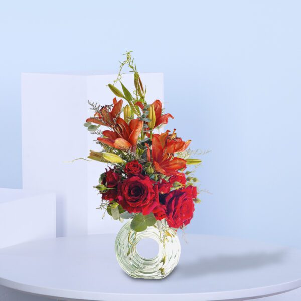 A stunning Valentine's Day floral arrangement with red roses, orange lilies, and lush greenery in a clear glass vase.