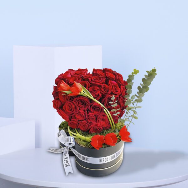 Round flower box with a romantic heart-shaped arrangement of red roses with green accents.