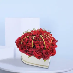 The image shows a beautiful arrangement of red roses in a heart-shaped box. The roses are arranged in a way that creates a full and lush look, with some of the roses even spilling over the edges of the box