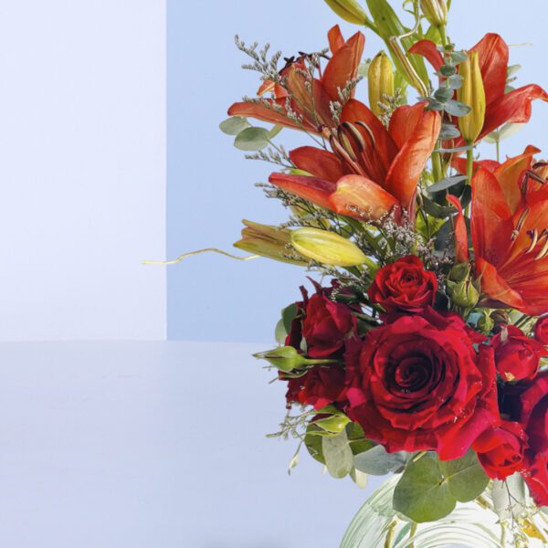 A stunning Valentine's Day floral arrangement with red roses, orange lilies, and lush greenery in a clear glass vase.