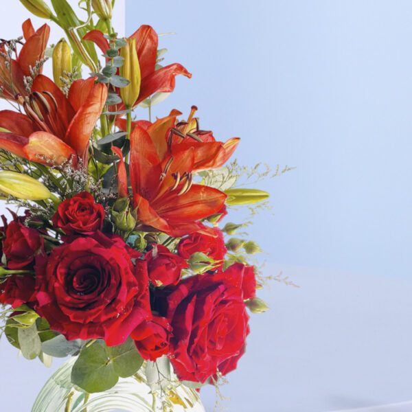 A stunning Valentine's Day floral arrangement with red roses, orange lilies, and lush greenery in a clear glass vase.