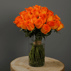 A bouquet of orange roses in a clear glass vase sits on a wooden surface against a gray background.